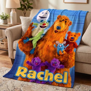 May include: A blue fleece blanket featuring characters from Bear in the Big House, including a large orange bear, a green lemur, a small blue mouse, two purple otters, and a moon, with the name Rachel printed in large yellow letters at the bottom.