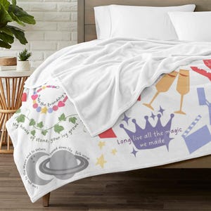May include: White plush blanket with colourful illustrations and text. The blanket features a crown, champagne glasses, and a planet with a crescent moon. Text includes "Long live all the magic we made" and other phrases.