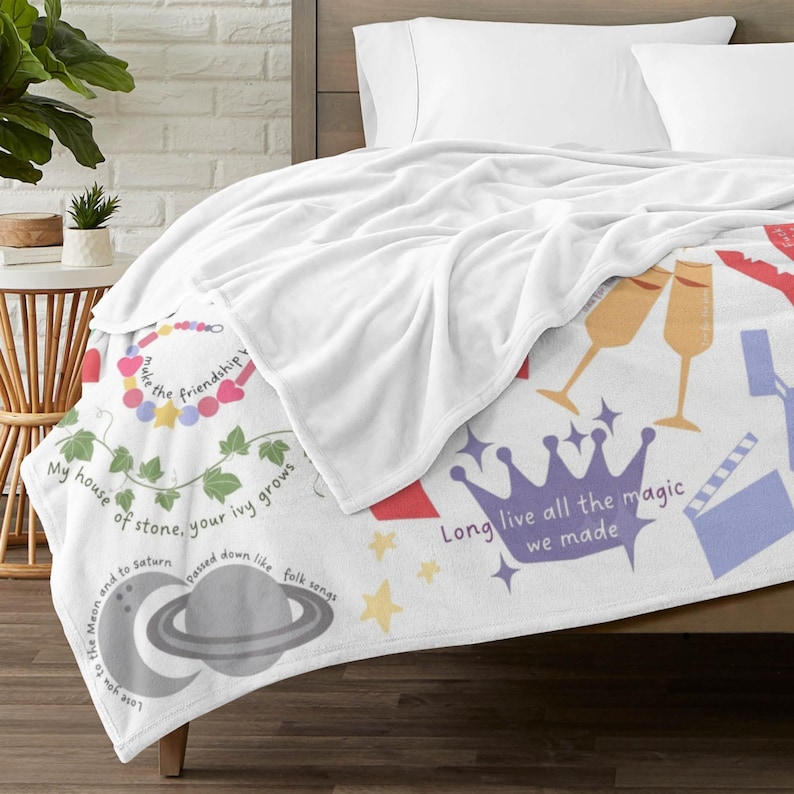May include: White plush blanket with colourful illustrations and text. The blanket features a crown, champagne glasses, and a planet with a crescent moon. Text includes "Long live all the magic we made" and other phrases.