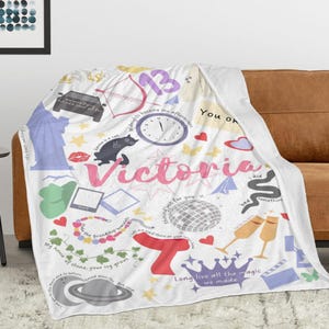 May include: White fleece blanket with a colourful design featuring the name "Victoria" in pink, along with illustrations like a clock, cat, and crown. The blanket has text elements such as "Long live all the magic we made."