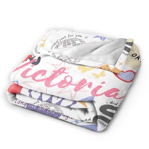 May include: A folded white blanket with the name "Victoria" in pink script. The blanket features a pattern of stars, butterflies, and phrases like "shining just for you" and "Long live all the magic".