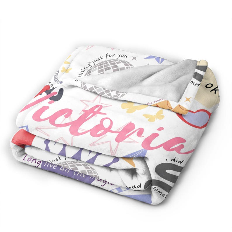 May include: A folded white blanket with the name "Victoria" in pink script. The blanket features a pattern of stars, butterflies, and phrases like "shining just for you" and "Long live all the magic".