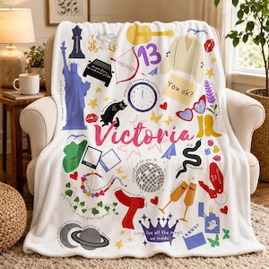 May include: White fleece blanket with colorful illustrations, including a guitar, a clock, a car, and the name "Victoria" in pink. The blanket has various text elements.
