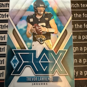 May include: A football trading card featuring Trevor Lawrence of the Jacksonville Jaguars. The card has a teal and silver design with the player in uniform holding a football. The card's logo reads "REX" with the player's name and team at the bottom.
