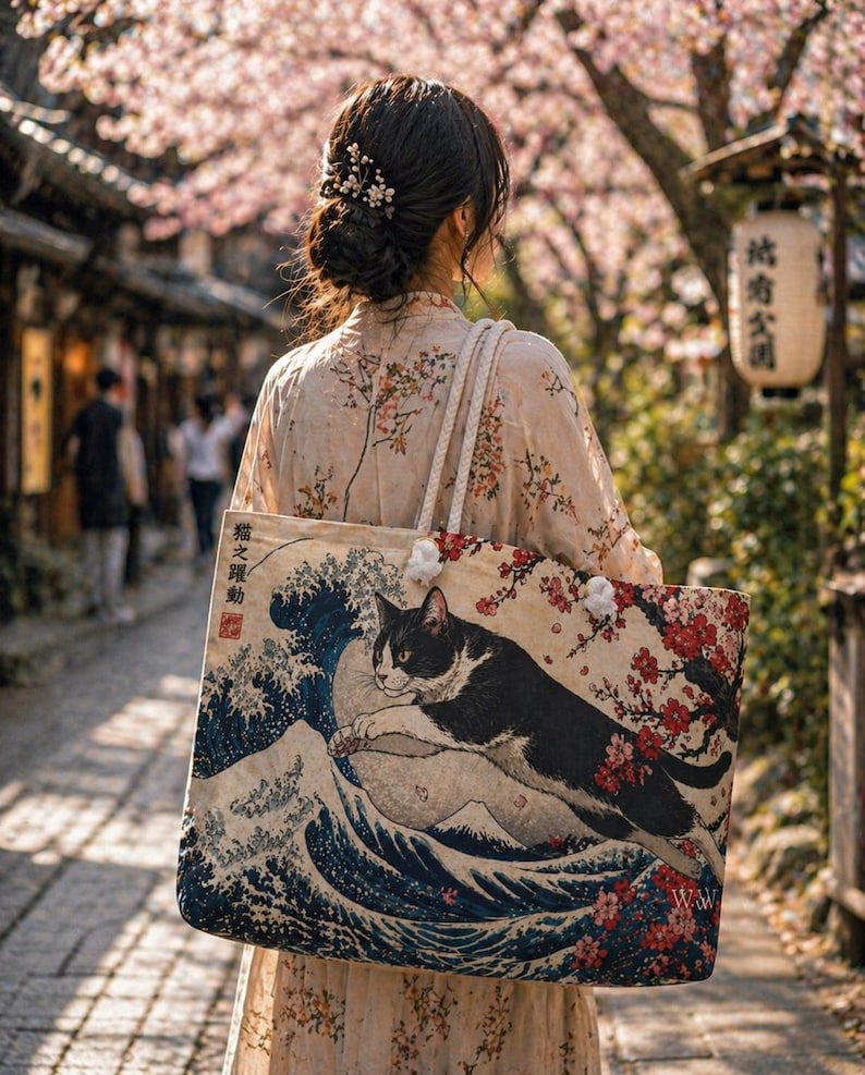 May include: A beige tote bag featuring a black and white cat leaping over a wave, inspired by Japanese art. The bag has a floral design and white straps. The image also shows a person wearing a floral patterned dress.