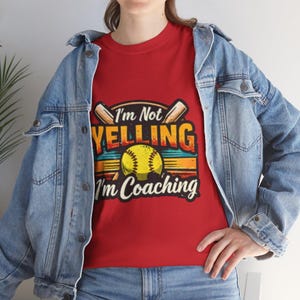 Könnte beinhalten: Rotes T-Shirt mit dem Text "I'm Not Yelling I'm Coaching" in einem farbenfrohen Design mit einem Softball und Schlägern. Das Shirt wird mit einer blauen Jeansjacke getragen.