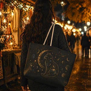 May include: A black tote bag with a celestial cat design. The bag features a gold crescent moon, stars, and planets. The tote bag has white rope handles and is being carried by a person wearing a dark gray sweater.