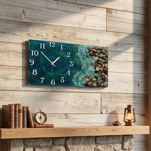 May include: A rectangular wall clock featuring a deep teal and blue ocean wave design with white foam accents and a cluster of smooth brown river stones on the right side. The clock face displays white Arabic numerals and thin white clock hands.
