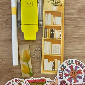May include: A yellow portable reading light, a white pen, a tall bookmark with a bookshelf illustration, a tulip bookmark, a silver paperclip, and four stickers featuring books, flowers, and the text Drink Tea Read Books Be Happy and bookish.