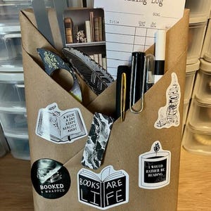 May include: A brown paper-wrapped package with stickers and stationery. Stickers include book-themed quotes and images. Inside are pens, a reading log, and decorative feathers. The package is on a wooden surface.