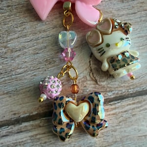 May include: A charm necklace featuring a pink bow, a Hello Kitty charm, and other decorative elements. The Hello Kitty charm has a leopard print dress. Other charms include a pink beaded ball, a heart-shaped bead, and a bow with a leopard print design.