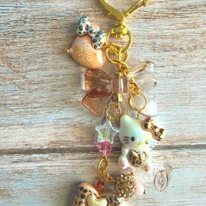May include: A gold-colored keychain with multiple charms. Charms include a Hello Kitty figure, bows, hearts, stars, and beads. The keychain has a heart-shaped clasp and a mix of leopard print and pastel colors.