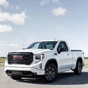 Könnte beinhalten: Ein weißer GMC Sierra Pickup-Truck mit schwarzem Kühlergrill und roten GMC-Schriftzügen. Der Truck hat schwarze Räder und ist auf einer gepflasterten Oberfläche unter blauem Himmel mit Wolken geparkt.