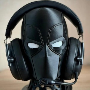 May include: A black headphone stand shaped like a superhero mask, with white eye cutouts. The stand holds a pair of black headphones with a microphone. The stand is on a wooden surface.
