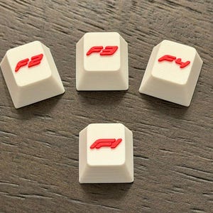 May include: Four white keyboard keycaps with red F1 logos and numbers. The keycaps are arranged on a dark wood-grain surface. The keycaps have the numbers F2, F3, F4, and the F1 logo.