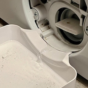 Op de afbeelding: Close-up van een witte wasmachine met het filtercompartiment open. Een witte bak is onder de opening geplaatst en vangt water op. Het filter is zichtbaar in de machine en water stroomt in de bak.