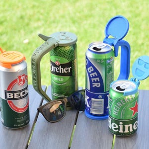 May include: An assortment of beer cans with colorful can toppers and a pair of sunglasses on a wooden table. The cans include Beck's, Dreher Gold, and Heineken. The can toppers are orange, blue, and green.