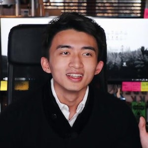May include: A person wearing a black jacket over a white shirt, sitting in front of multiple computer monitors. The monitors display various images and have sticky notes attached. A keyboard and other equipment are visible.