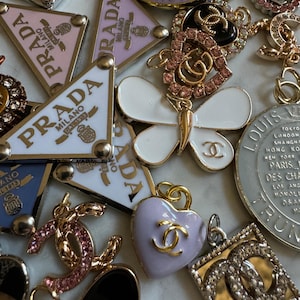May include: A collection of designer charms and pendants in various shapes and colors. Includes Prada triangle charms, a white butterfly pendant, a heart-shaped charm with a Chanel logo, and other luxury brand emblems. Gold and silver tones are prominent.