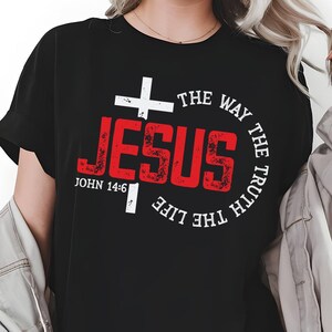 Puede incluir: Camiseta negra con una cruz blanca y la palabra "JESUS" en letras rojas desgastadas. El texto "THE WAY THE TRUTH THE LIFE" está en un diseño circular alrededor de la palabra "JESUS". La referencia bíblica "JUAN 14:6" también es visible.