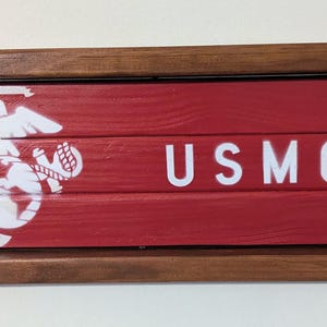 May include: A rectangular wooden wall sign featuring a dark brown stained frame and three horizontal red wooden slats. The sign displays a white United States Marine Corps eagle, globe, and anchor emblem on the left and the white text USMC on the right.