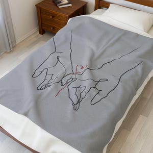 May include: A gray blanket featuring a minimalist line drawing of two hands reaching for each other, connected by a red thread. The blanket is on a bed with a white sheet and pillow. The design is simple and elegant.
