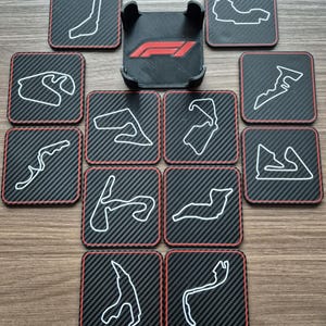 Può includere: Un set di sottobicchieri quadrati con texture in fibra di carbonio e bordi rossi, ognuno con il contorno bianco di un circuito di gara. Un supporto per telefono nero con il logo F1 al centro. I sottobicchieri sono disposti attorno.