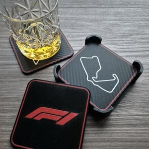 Könnte beinhalten: Set aus drei quadratischen Untersetzern mit schwarzem Karbonfaser-Design und rotem Rand. Ein Untersetzer zeigt das Formel-1-Logo in Rot, ein weiterer eine Rennstreckenkontur, und der dritte trägt ein Glas mit bernsteinfarbener Flüssigkeit.