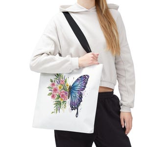 May include: White tote bag featuring a watercolor design of a butterfly with blue and purple wings, and a bouquet of pink and yellow roses. The bag has a black shoulder strap.