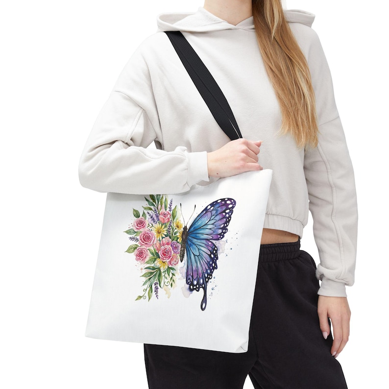 May include: White tote bag featuring a watercolor design of a butterfly with blue and purple wings, and a bouquet of pink and yellow roses. The bag has a black shoulder strap.