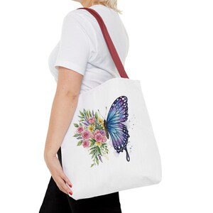 May include: White tote bag with a burgundy strap featuring a watercolor design of a blue and purple butterfly and a bouquet of pink and yellow flowers. The bag is carried over the shoulder.