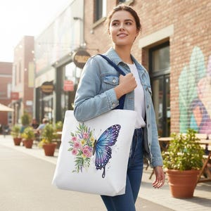 May include: White tote bag featuring a watercolor design of a blue and purple butterfly with a bouquet of pink and yellow flowers. The bag has a navy blue strap and is carried by a person.