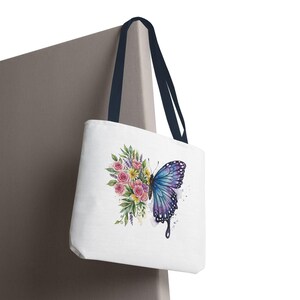 May include: White tote bag with a watercolor design of a butterfly and flowers. The butterfly is blue and purple, and the flowers are pink, yellow, and green. The bag has navy blue handles.