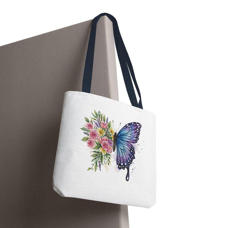 May include: White tote bag with a watercolor design of a butterfly and flowers. The butterfly is blue and purple, and the flowers are pink, yellow, and green. The bag has navy blue handles.
