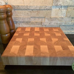 May include: A rectangular end-grain cutting board with a checkerboard pattern, made of light and dark wood. Two wooden salt and pepper grinders stand to the left. The board is approximately 12 inches wide and 18 inches long. A green and white logo is in the bottom right corner.