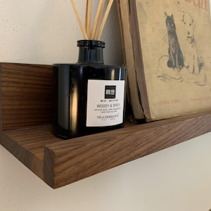 May include: A dark wooden shelf displays a black diffuser bottle with reeds, a vintage book with dog illustrations, and a black candle. The diffuser bottle has a white label with text. The shelf is mounted on a white wall.