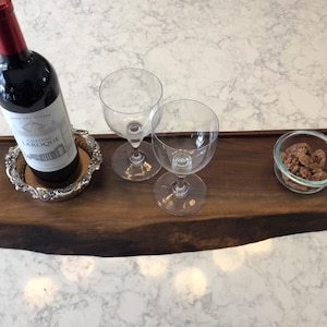 Puede incluir: Una bandeja de madera con borde natural, que contiene una botella de vino tinto, dos copas de vino y un cuenco pequeño con aperitivos. La bandeja tiene asas de latón y un posavasos de plata.