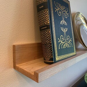 May include: A wooden floating shelf with a light finish, mounted on a beige wall. A hardcover book, a decorative plate, and a black diffuser bottle are displayed on the shelf. The book has gold accents and the plate has a gold rim.
