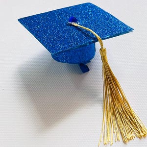 May include: A miniature graduation cap featuring a vibrant blue glitter finish on the square mortarboard and base. A small, dark blue gem is centered on top, attached to a long, metallic gold tassel that hangs down from the center point.