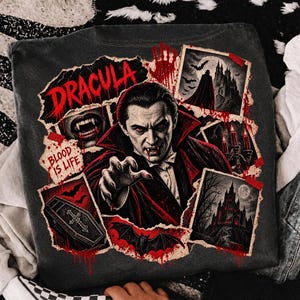 Puede incluir: Una camiseta gris carbón con un estampado gráfico de Drácula con colmillos, una capa forrada en rojo y una mano extendida. El diseño incluye el texto "DRACULA" y "BLOOD IS LIFE" junto a imágenes de un castillo, un ataúd y murciélagos.