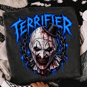 May include: A charcoal grey t-shirt featuring a graphic print of a clown face with a small black top hat, blood splatters, and a surrounding metal chain. The word TERRIFIER is printed in a bright blue, jagged, dripping font above the clown.