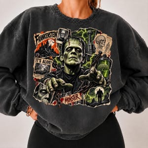 May include: A charcoal gray crewneck sweatshirt featuring a large graphic print of Frankenstein's monster. The design includes the text "FRANKENSTEIN", "IT'S ALIVE!", and "MONSTER", alongside imagery of a castle, a brain in a jar, and lightning.