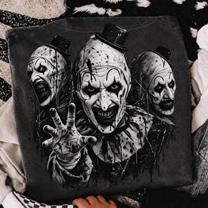 May include: A dark gray t-shirt featuring a graphic print of three identical clowns wearing top hats. The central clown is reaching forward with an outstretched hand. The design is rendered in high-contrast black and white with splatter details.