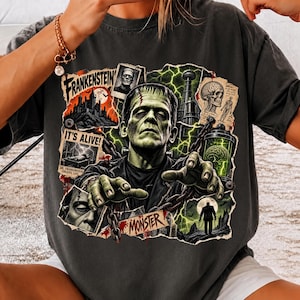 May include: A charcoal gray t-shirt featuring a graphic print of Frankenstein's monster with green skin, surrounded by collage-style imagery including a castle, a skull, a brain in a jar, chains, and text reading Frankenstein, It's Alive, and Monster.