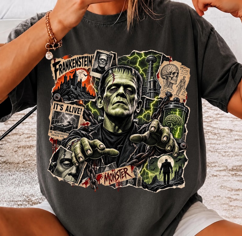 May include: A charcoal gray t-shirt featuring a graphic print of Frankenstein's monster with green skin, surrounded by collage-style imagery including a castle, a skull, a brain in a jar, chains, and text reading Frankenstein, It's Alive, and Monster.