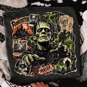 May include: A charcoal gray t-shirt featuring a graphic print of Frankenstein's monster with outstretched hands and chains. The design includes text reading Frankenstein, It's Alive, and Monster, alongside imagery of a castle, a brain in a jar, and a skull.
