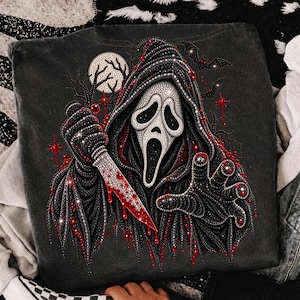 May include: A black t-shirt featuring a rhinestone-embellished graphic of a hooded figure with a white mask, holding a knife with red accents. The design includes a full moon, bare tree branches, and flying bats, all rendered in sparkling crystals.