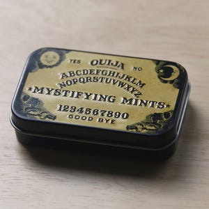 May include: A rectangular black metal tin featuring a gold-colored lid with a Ouija board design. The lid displays the alphabet, numbers zero through nine, the words YES, NO, GOOD BYE, and the text MYSTIFYING MINTS, along with sun and moon graphics.