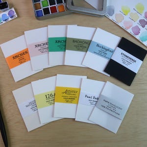 May include: A collection of eleven small rectangular watercolor paper samples with brand labels including Arches, Baohong, Bockingford, Stonehenge, Canson 1264, Fabriano Artistico, Paul Rubens, and Yupo. A black paintbrush and a metal watercolor paint tin are included.