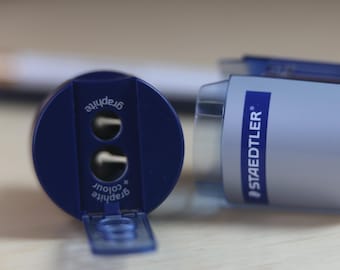 May include: A blue and grey Staedtler pencil sharpener with two holes for different pencil sizes. The sharpener features a hinged lid and is labeled with the text graphite and colour on the front, with the brand name printed on the side.